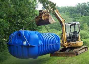 A backhoe lifts a blue septic tank off the ground.