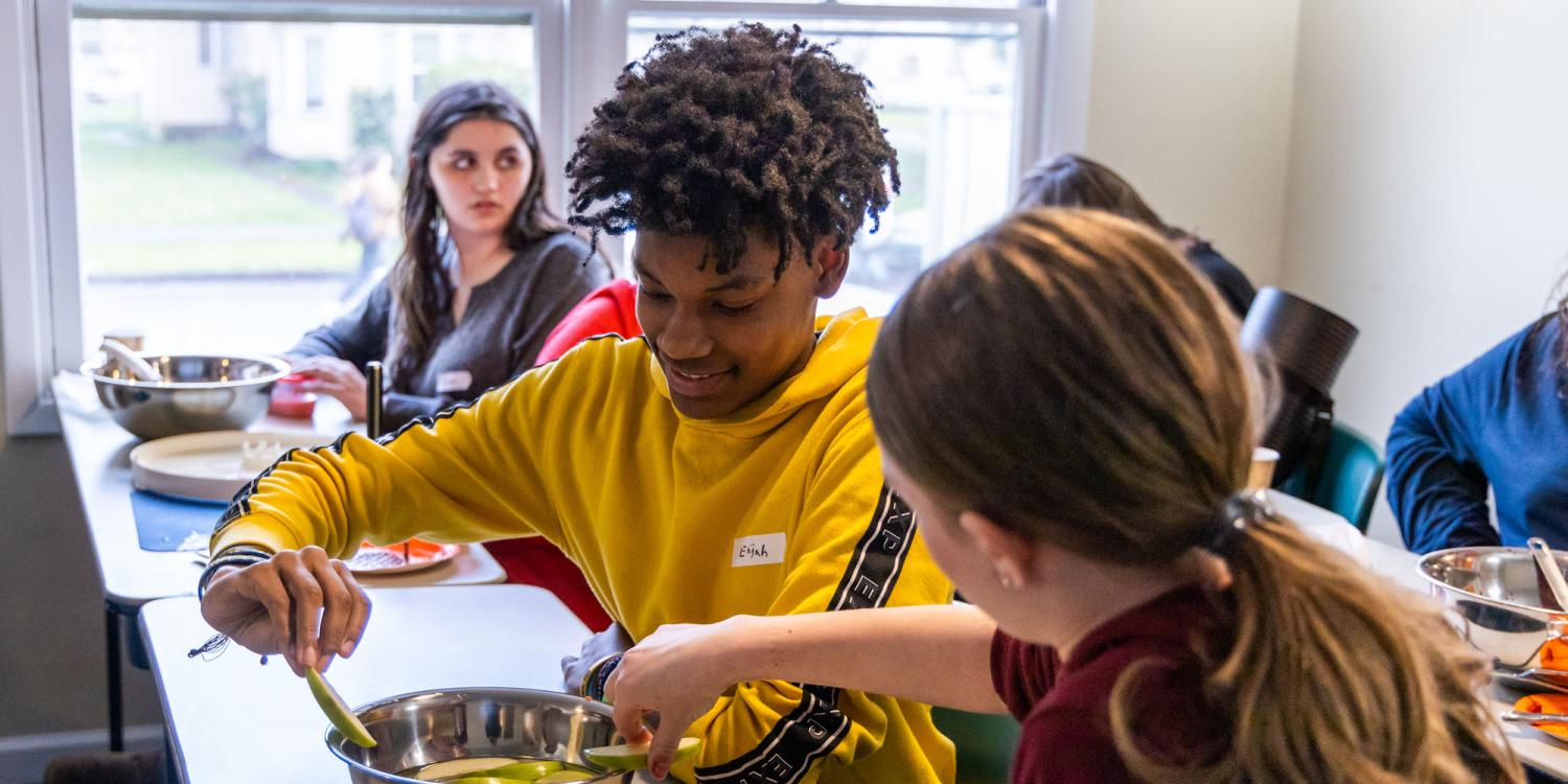 Two young people work together slicing cucumbers into a metal bowl during a food preparation activity.