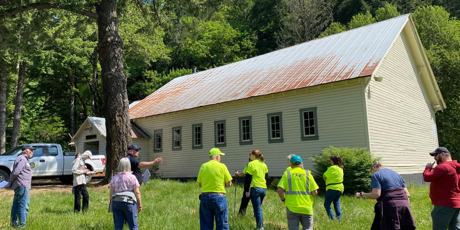 A group of people, some in bright safety vests, stand outside a historic-looking building in a forested area, conducting a home ignition zone assessment.