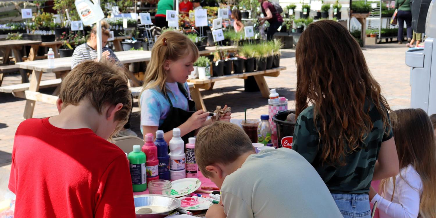 Children gather around a table filled with art supplies, painting and crafting together at an outdoor event surrounded by plants and picnic tables.