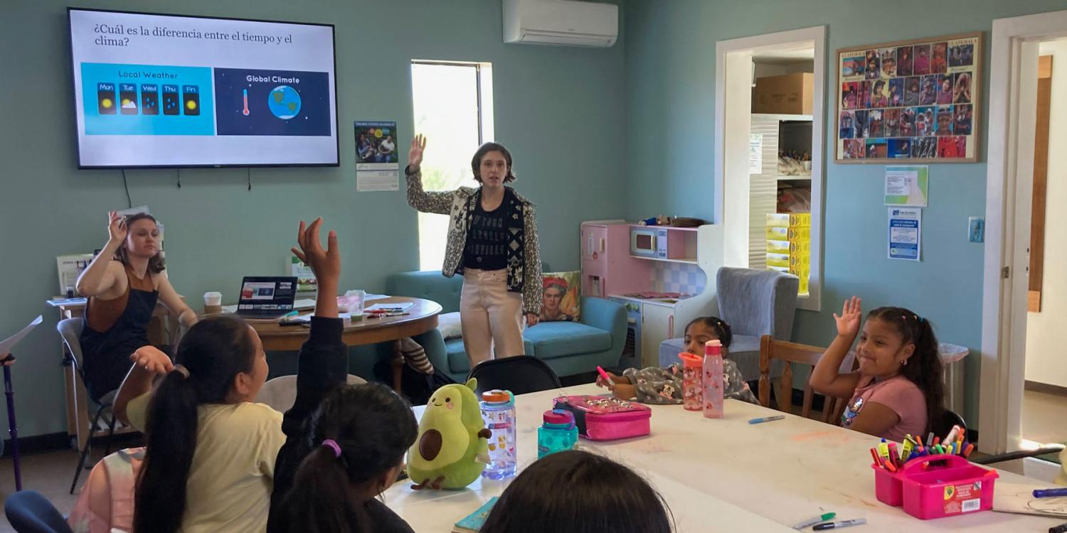 A group of children and two instructors are engaged in a classroom activity about weather and climate, with several students raising their hands to participate.