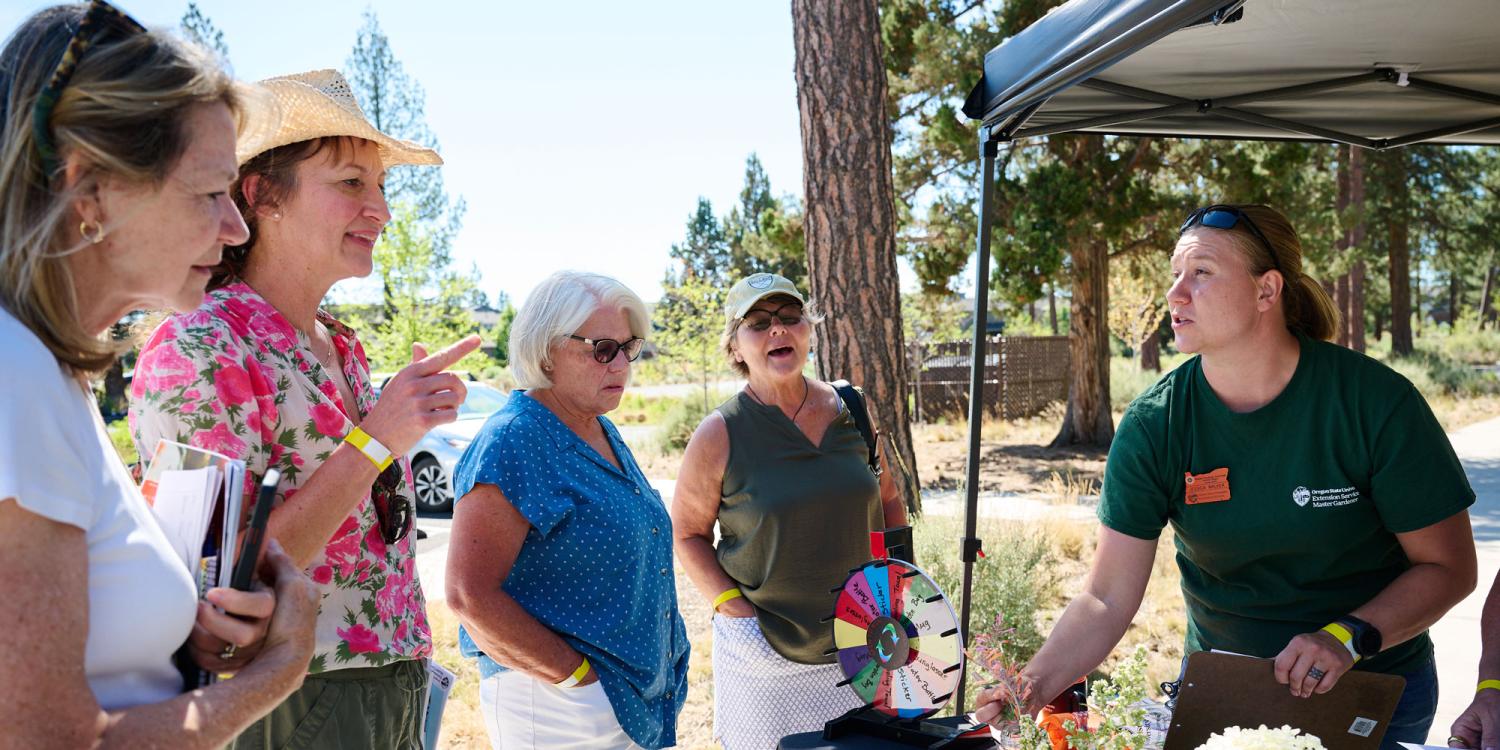 OSU Extension Master Gardener volunteer talks with a small group of adults at an outdoor booth during the 2025 High Desert Garden Tour, using a display and materials to answer questions.