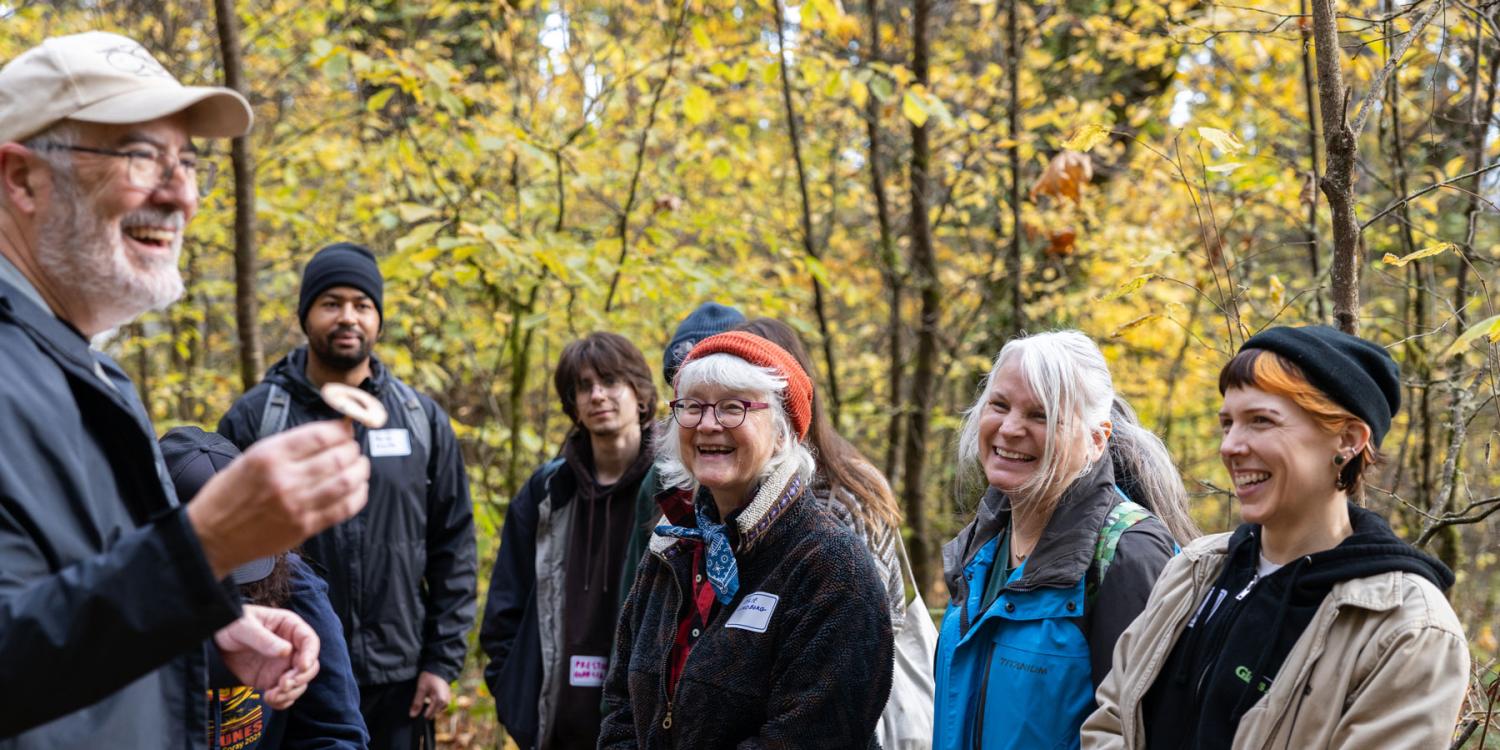 A group of adults listen and smile as a man holds up a mushroom during a guided walk in a wooded area with fall foliage.