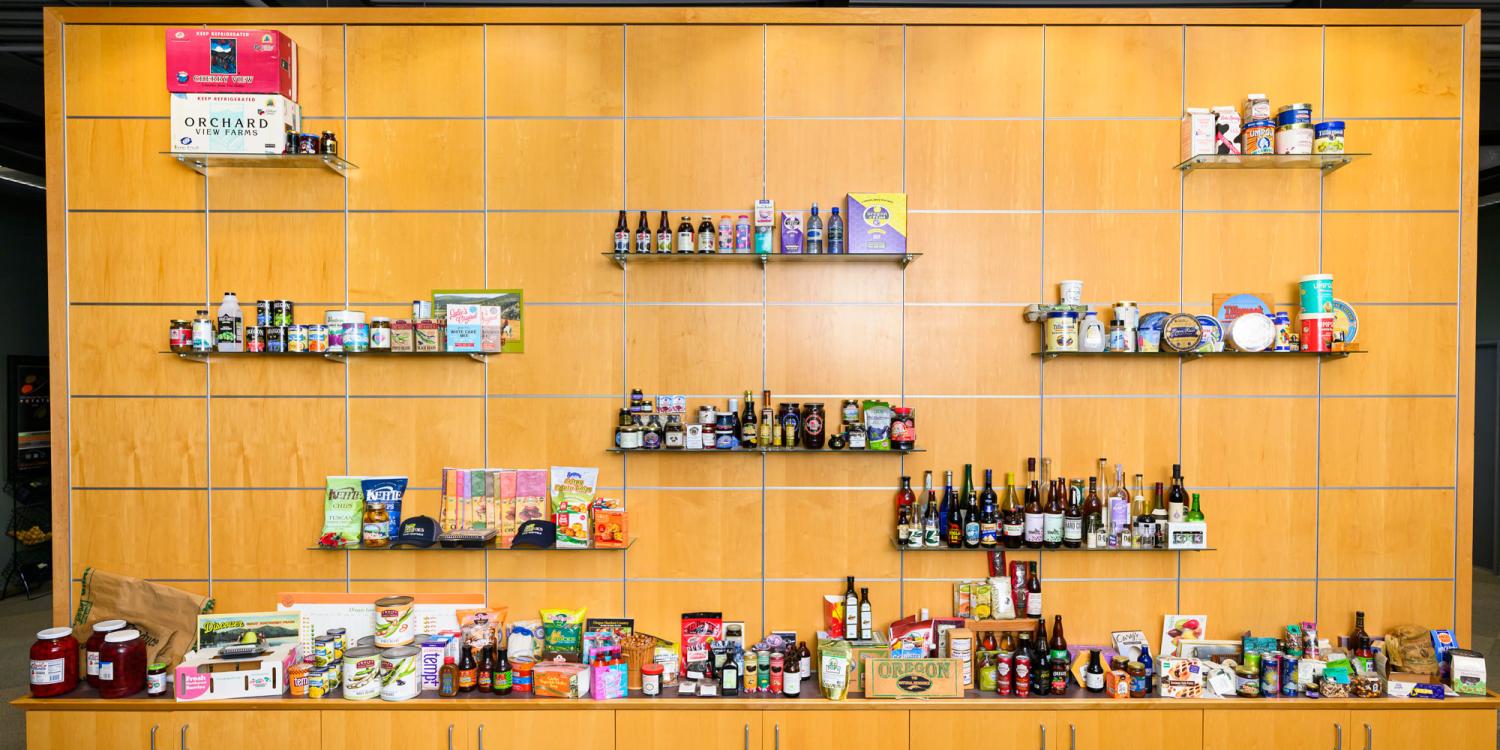 A large display wall features shelves of Oregon-made food and beverage products, including packaged foods, jars, bottles and boxes arranged across a wood-paneled backdrop.