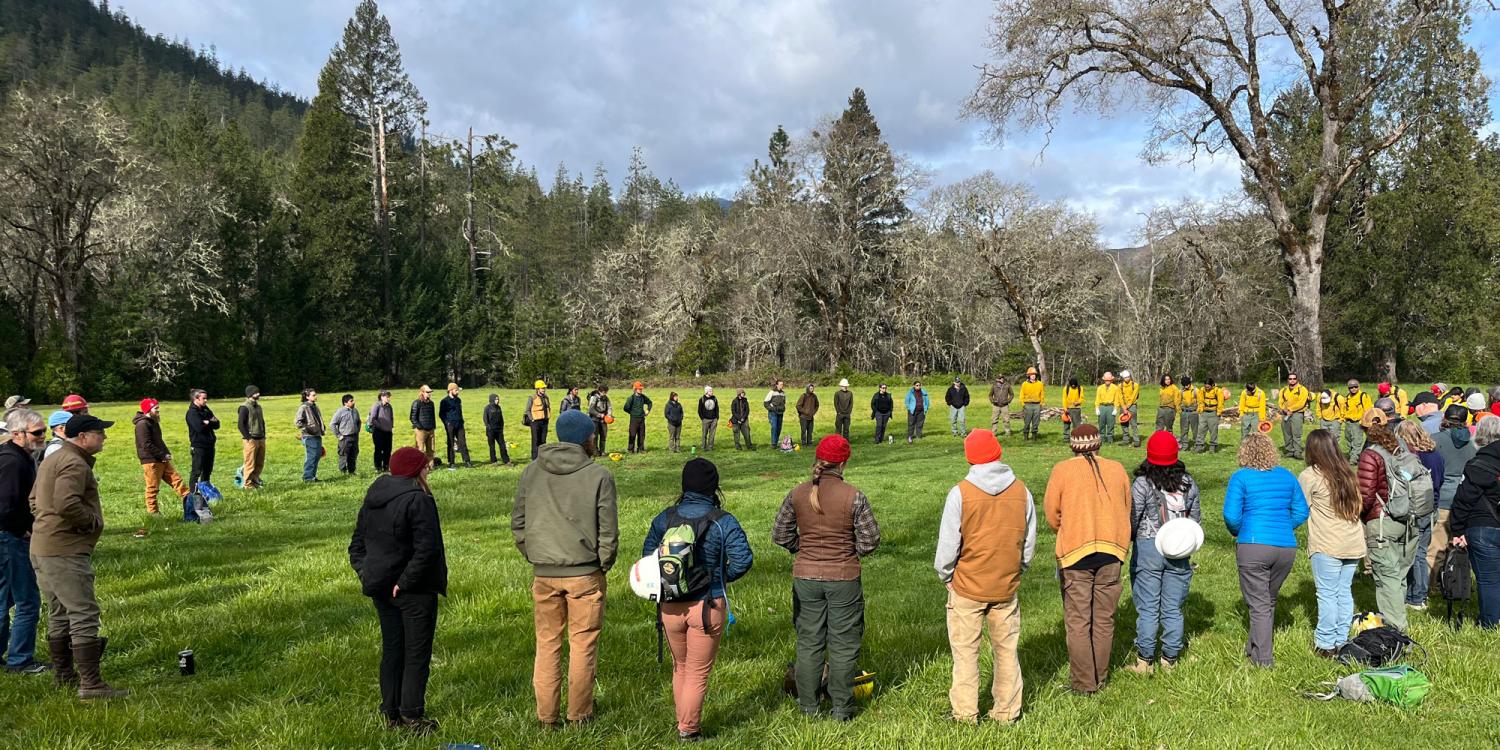 Participants and wildland fire personnel stand in a large circle in a grassy field surrounded by forested hills during an IGNITE prescribed fire skills training event.
