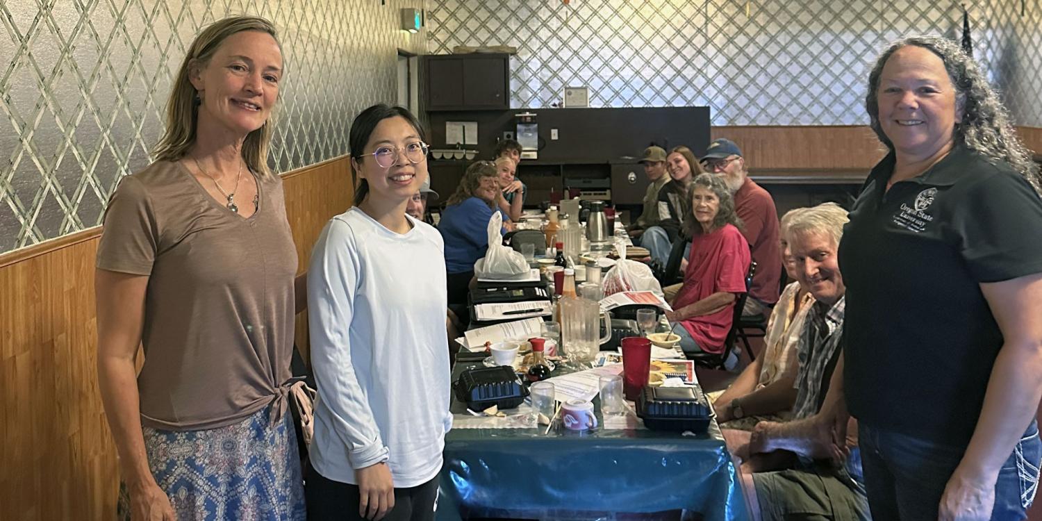 Members of the Josephine County Farm Bureau sit around a long restaurant table during a meeting while OSU Extension faculty Maud Powell, Nan Xu and Shelby Filley stand at the front of the group smiling for the camera.