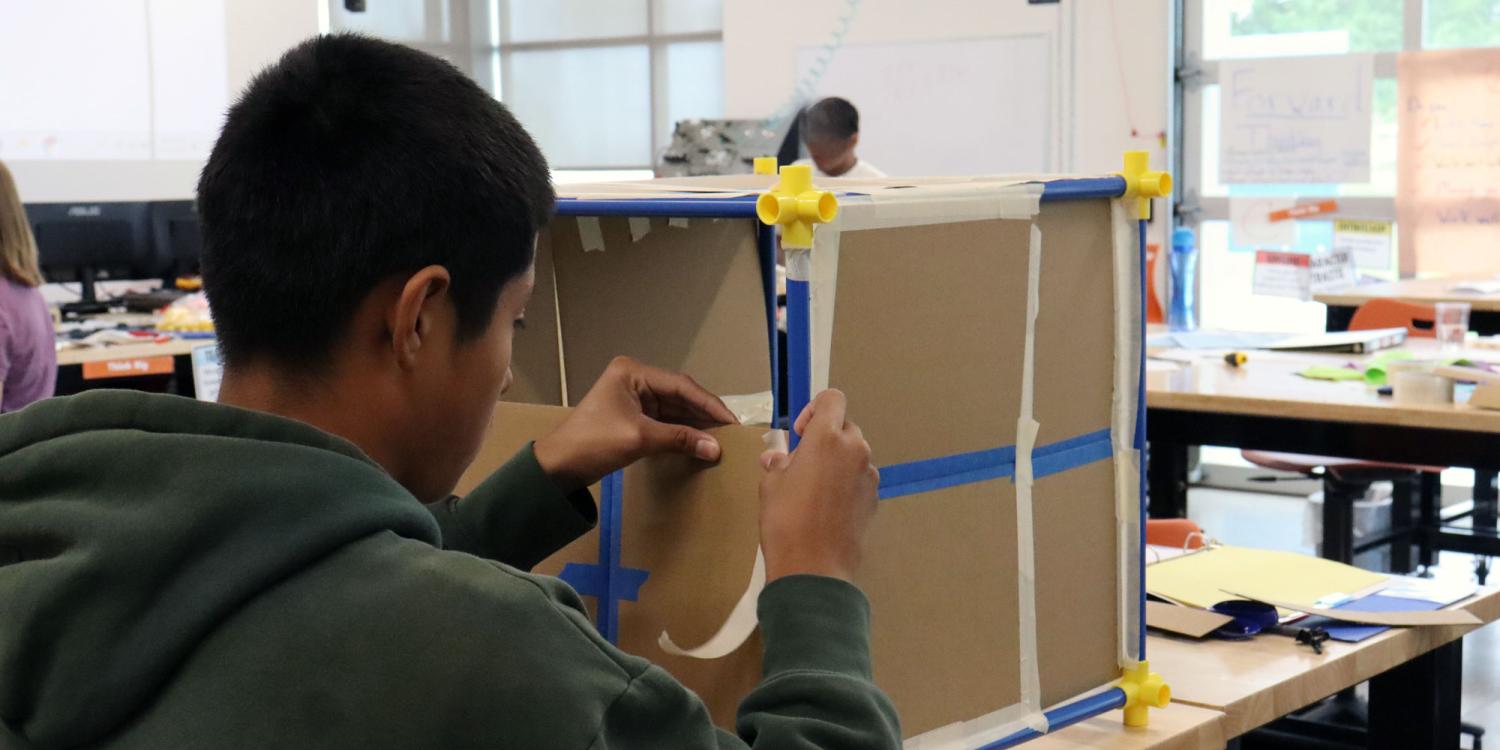 A youth works at a classroom table assembling a cardboard model structure during a hands-on career exploration activity.
