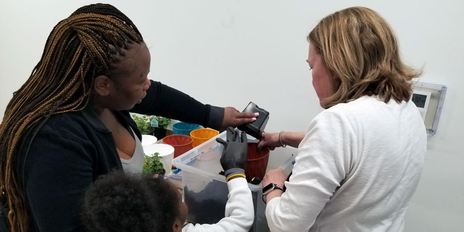 n adult and child pour potting soil into containers while another instructor helps during a hands-on Seed to Supper gardening activity indoors.