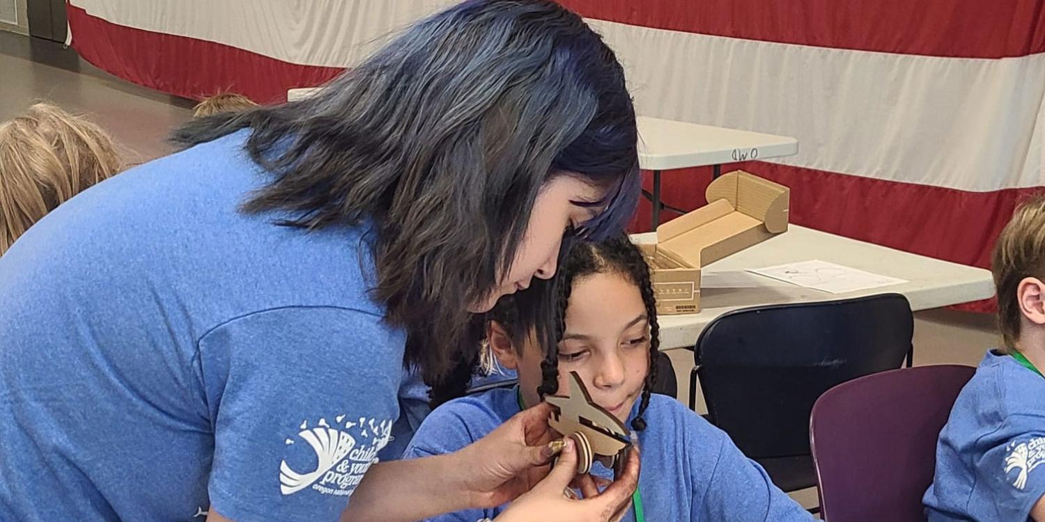 A teen mentor helps a child assemble a small wooden project during Military Kids Club camp, with other youths working nearby in front of a large American flag backdrop.