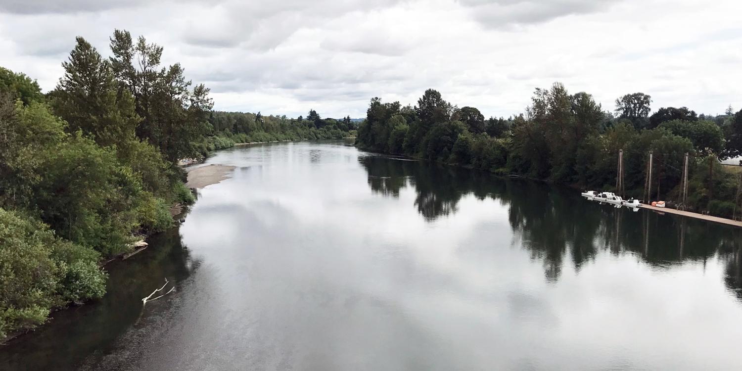 A calm river reflecting the cloudy sky is bordered by dense green trees on both sides, with a small dock visible to the right.