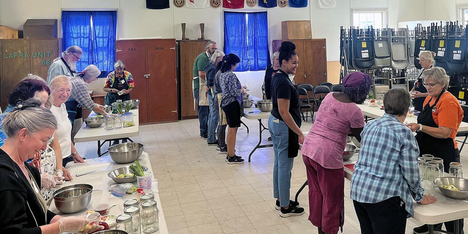 Participants work in small groups during a food preservation class, preparing ingredients and filling jars at tables set up throughout a community hall while instructors provide guidance.
