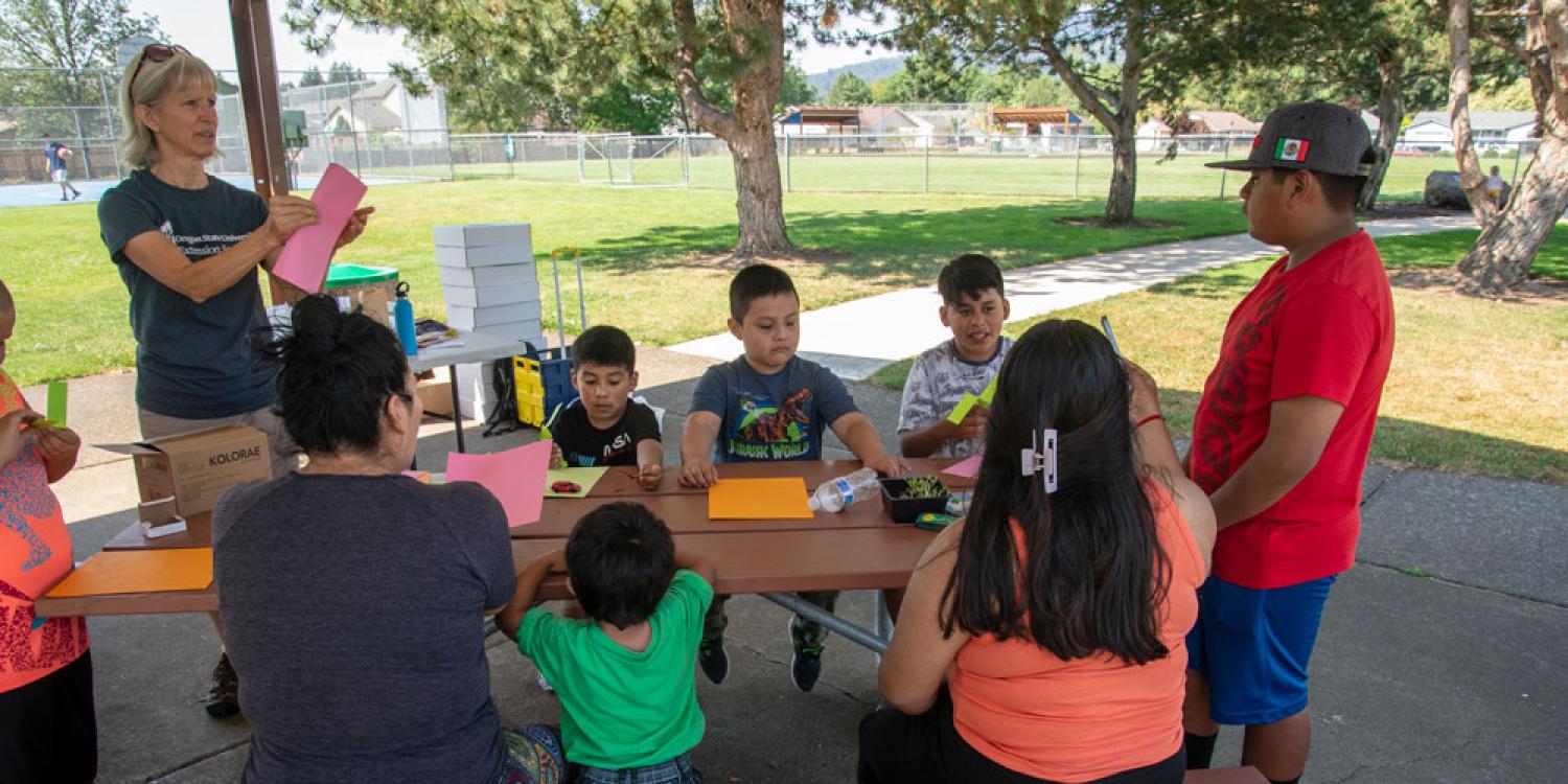 Latino children sit around a picnic table in a park making paper airplanes.