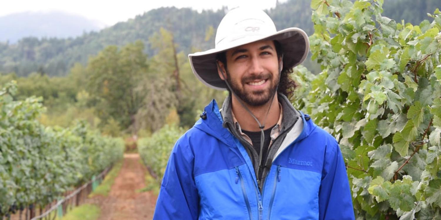 A man in a blue jacket and wide-brimmed hat stands smiling in a vineyard, surrounded by rows of grapevines and backed by forested hills.