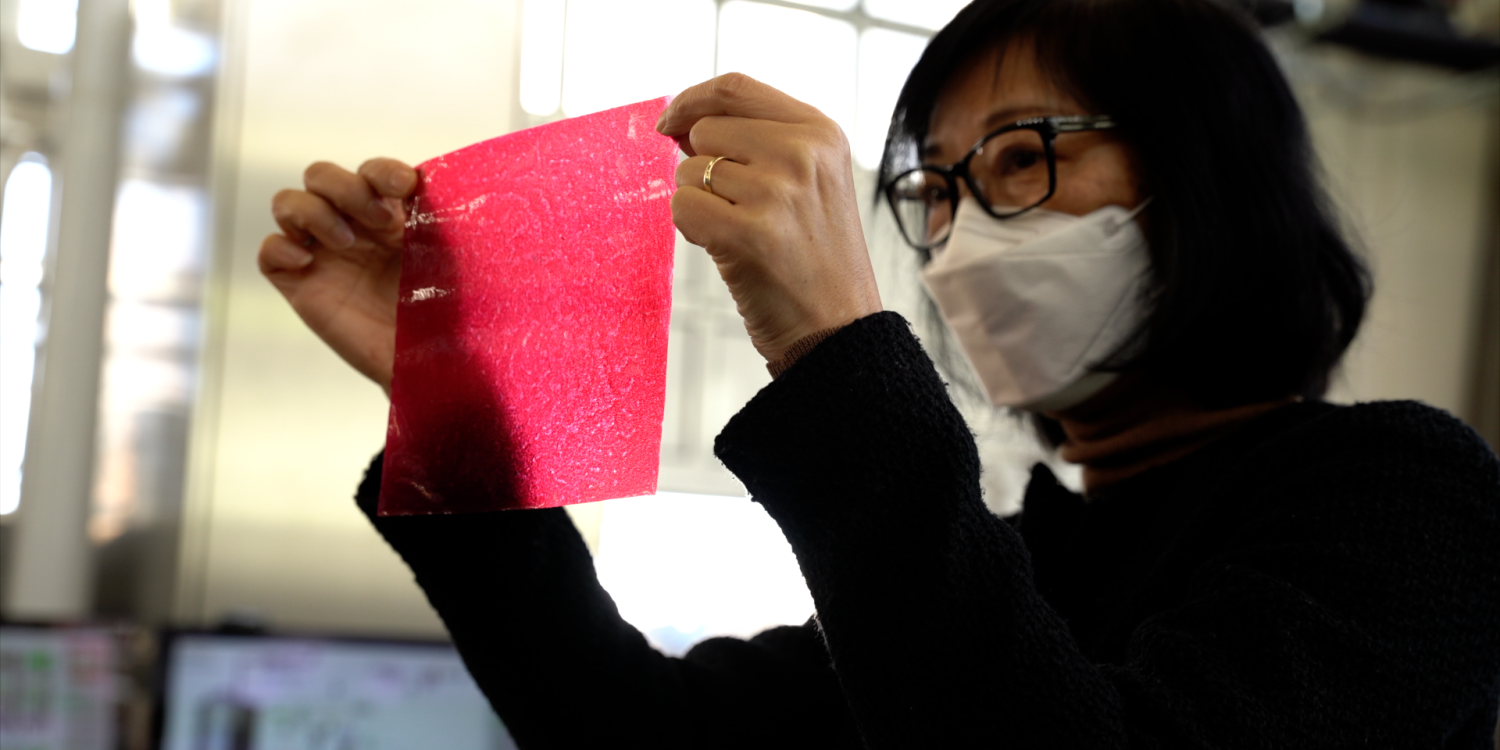 A woman holds up a thin, pink packaging material made of apple food waste.