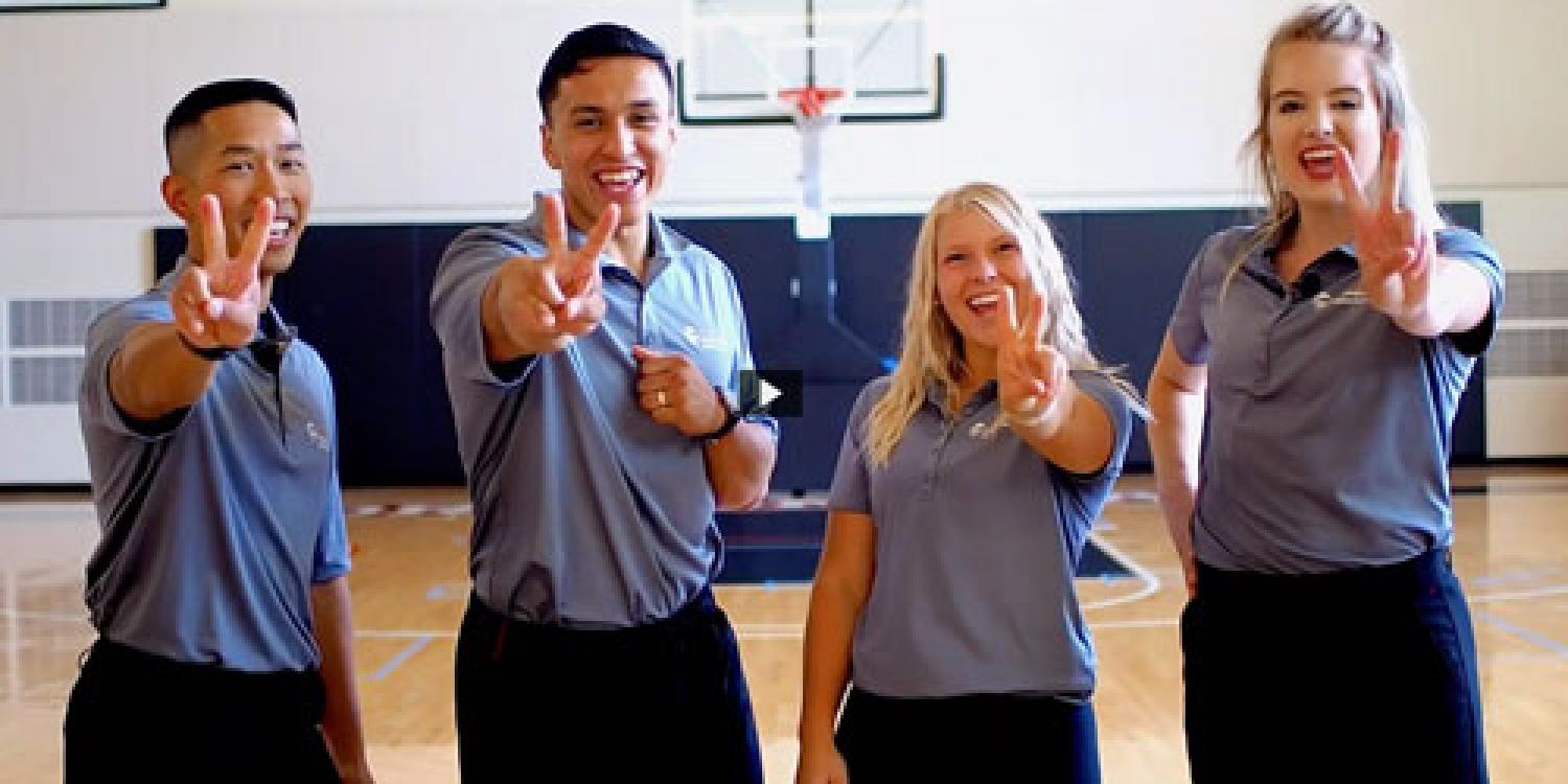 Four smiling young adults in matching gray polo shirts stand in a gym, holding up two fingers in front of a basketball hoop.