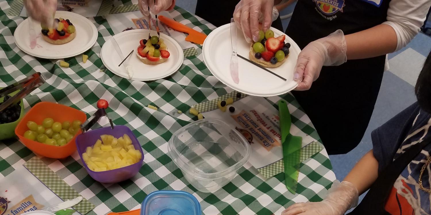 People wearing gloves assemble fruit pizzas on paper plates at a table covered with a green checkered tablecloth and containers of assorted fruit.