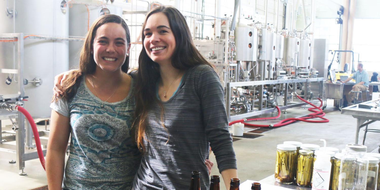Two young women pose in front of bottles of kombucha in a brewery.