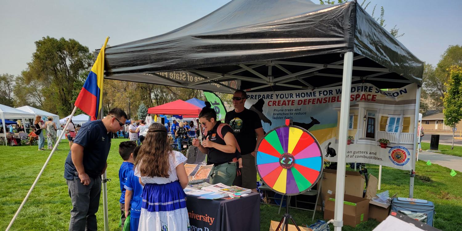 Sure! Here's a one-sentence alternative text description for the image:  "People gather at a community fair booth with wildfire preparedness materials, a colorful prize wheel, and a Colombian flag, while a woman in traditional dress talks to exhibitors."