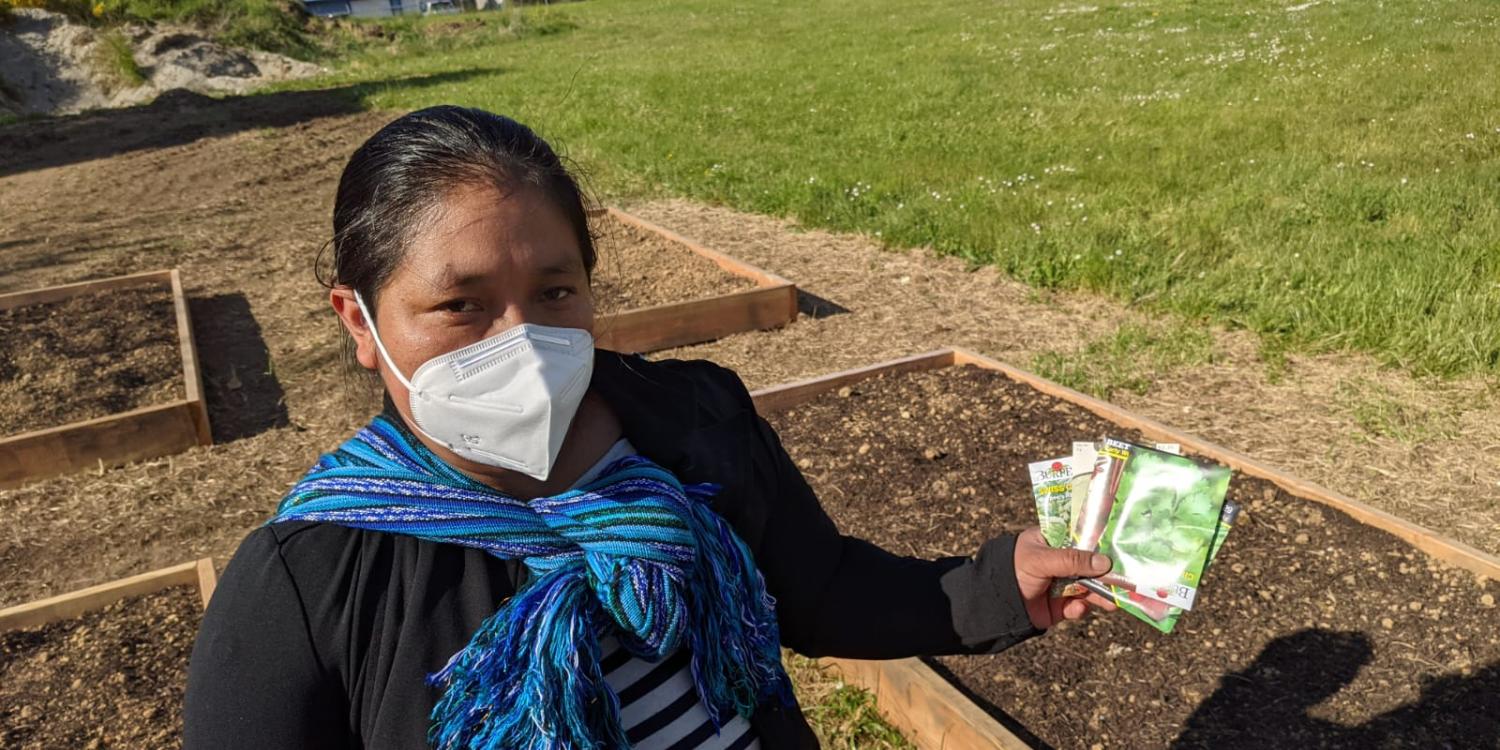 A community member wears a facemask and holds packets of seeds in her left hand. She is about to plant the seeds in a community garden in Lincoln County.