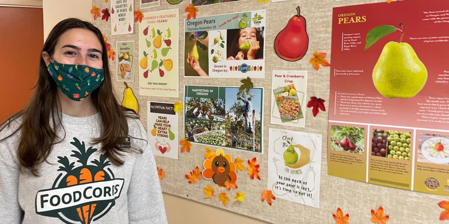 A young woman wearing a facemask stands next to a bulletin board in an elementary school hallway. She's wearing a sweatshirt that says FoodCorps on the front with a picture of carrots..