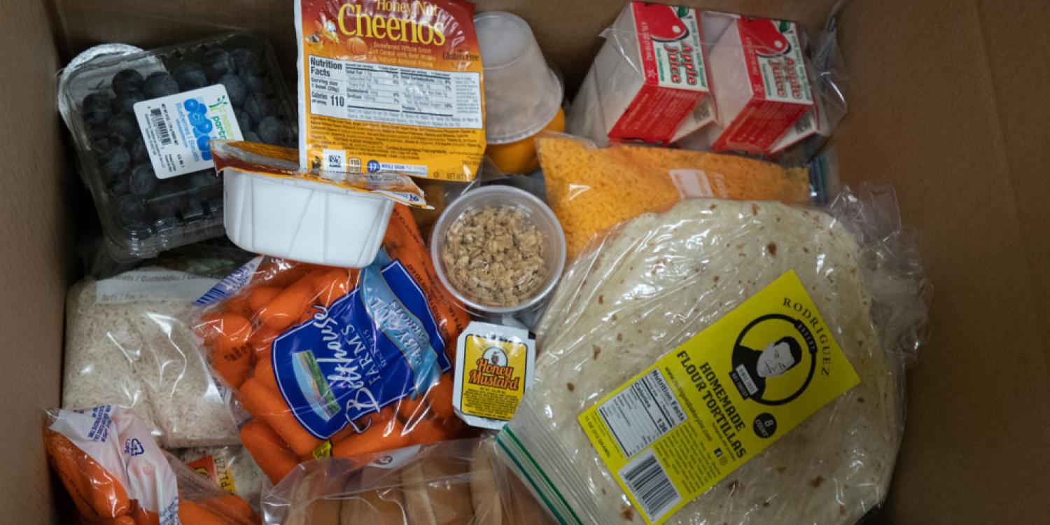 A box filled with food including tortillas, cereal, carrots, apple juice, rice, cheese, blueberries and bagels.