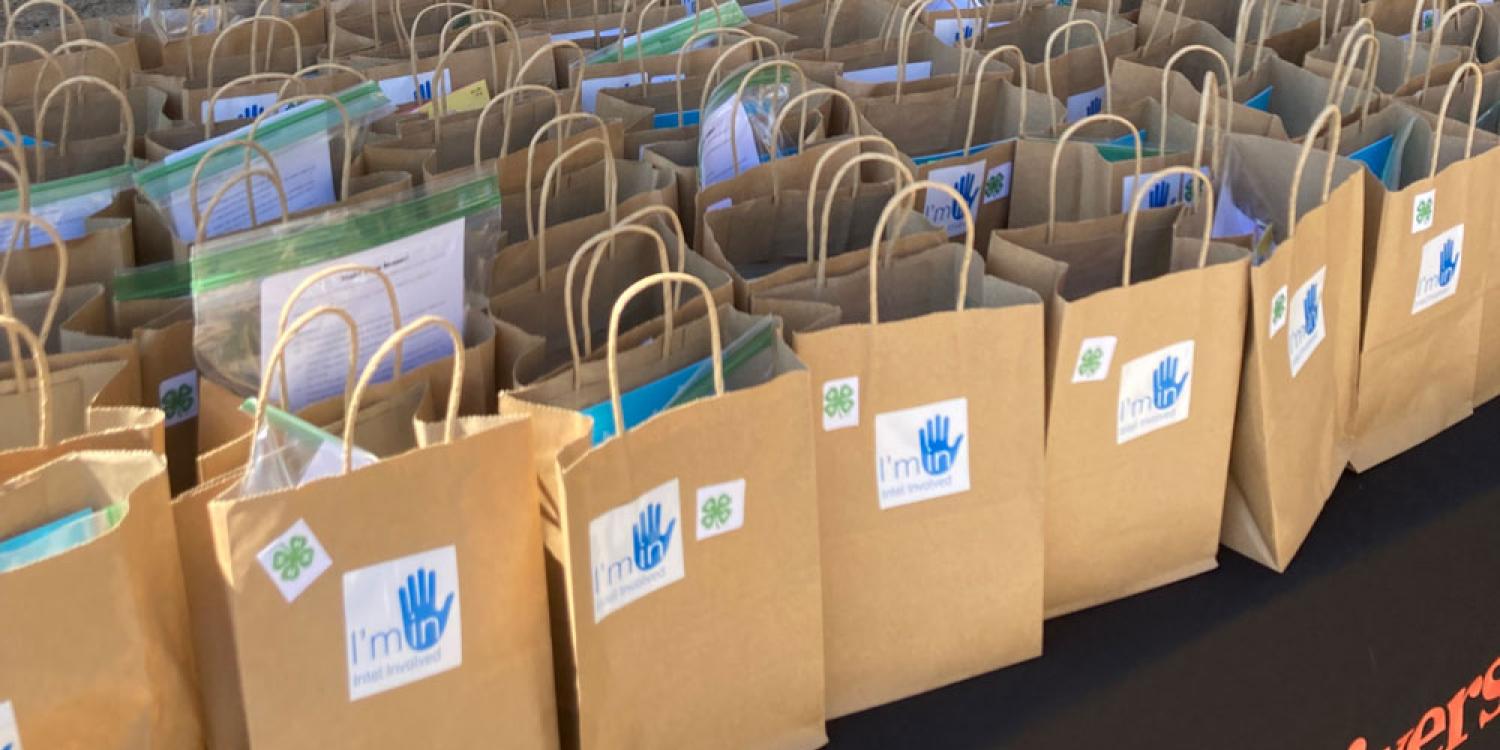 Brown paper bags, each filled with a pencil, eraser, snack item, Band-Aids, hand-written card, and a STEM kit, are lined up on a table.