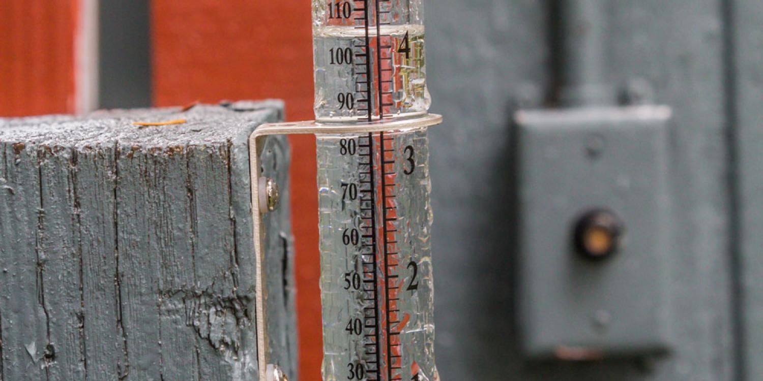 A close-up of a clear rain gauge mounted on a wooden post, showing a measurement of over 3 inches of collected rainfall.