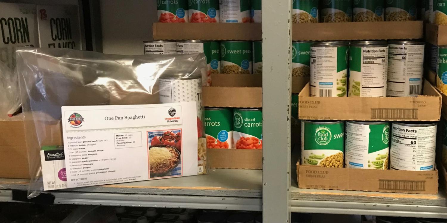 Shelves stocked with canned vegetables surround a plastic bag meal kit labeled “One Pot Spaghetti” that includes ingredients and a printed recipe.