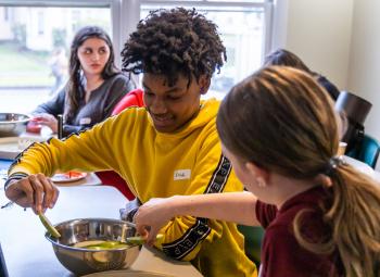 Two young people work together slicing cucumbers into a metal bowl during a food preparation activity.