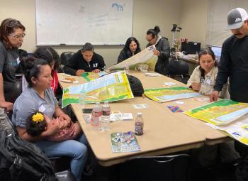 A group of adults and children gather around a table in a classroom, studying tsunami evacuation maps and emergency preparedness materials.