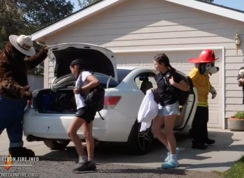 Two girls with backpacks exit a car parked in a driveway, greeted by mascots Smokey Bear and a fire dog character.
