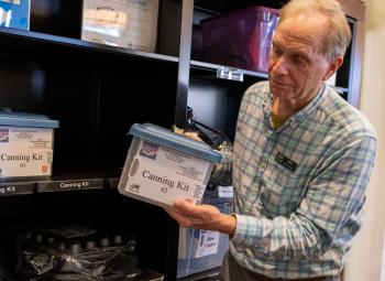 A man holds a labeled plastic container marked “Canning Kit” while standing in front of a shelf filled with similar educational kits in a library.