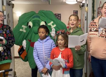 A group of people, including children and adults, sing holiday carols indoors while holding songbooks, with a person dressed in a green 4-H costume standing behind them.