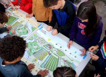 Children gather around a table, engaging with colorful illustrated farm maps and small cutout pieces in a hands-on educational activity.