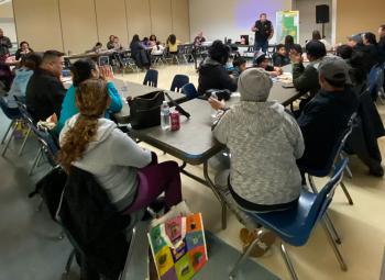 Community members sit at tables in a large room listening to a presenter speak in front of a projected screen and a safety map during an emergency preparedness event.