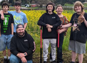 A group of smiling youth and an adult pose with tools in front of a field of yellow flowers during an agricultural education activity.