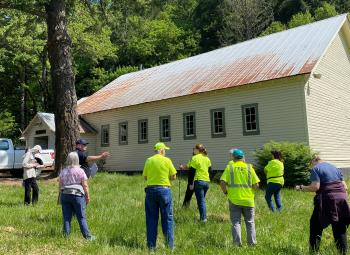 A group of people, some in bright safety vests, stand outside a historic-looking building in a forested area, conducting a home ignition zone assessment.