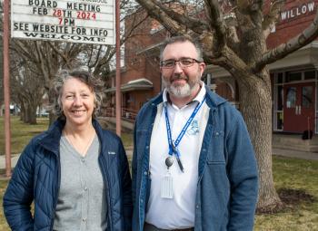 A woman and a man stand smiling outside a building with a sign announcing a February 2024 board meeting, surrounded by leafless trees and red-brick structures.