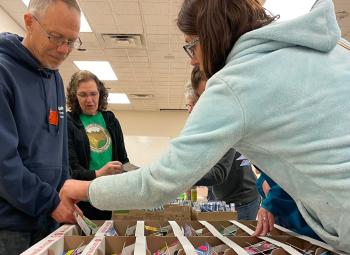 People browse and select seed packets from organized boxes during a Seed to Supper gardening event held indoors.