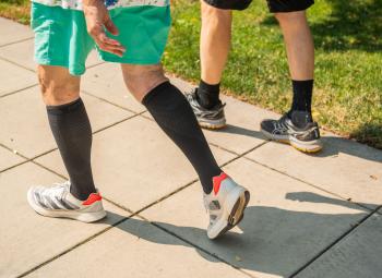 Two people walk along a sunny sidewalk, their legs visible as they wear athletic shoes and black compression socks.