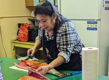 A woman wearing an apron prepares food at a kitchen counter, focused on cutting ingredients during a salmon canning session.