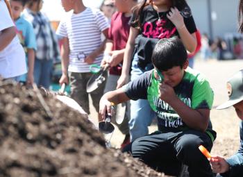 Children use magnifying glasses and trowels to explore a pile of soil during an outdoor agricultural education event.