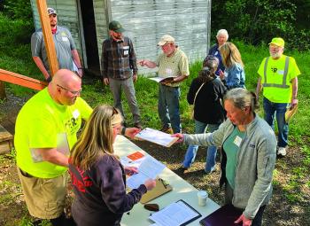 A group of adults participates in an outdoor emergency preparedness or training session, exchanging clipboards and instructions near a small building surrounded by greenery.