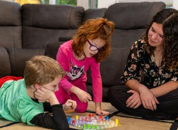 A young woman watches attentively as two children engage in an activity on the floor in a cozy living room.