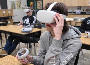 Students sit at desks in a classroom wearing virtual reality headsets and holding controllers, engaged in an immersive experience.