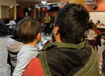 A father holds his young son while they watch a speaker address a community gathering in a decorated indoor space.