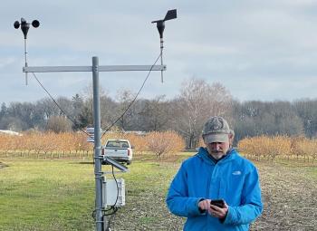 A man in a blue jacket checks his phone next to a weather station in an orchard field.