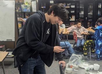A teenager looks at a phone while leading an activity in a classroom with younger students.