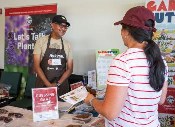 A Master Gardener volunteer wearing an “Ask me.” apron smiles and talks with a woman at an informational booth covered with seed displays and OSU Extension signs.