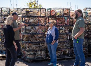 Four people stand talking in front of tall stacks of crab pots filled with ropes and buoys at a sunny harbor.